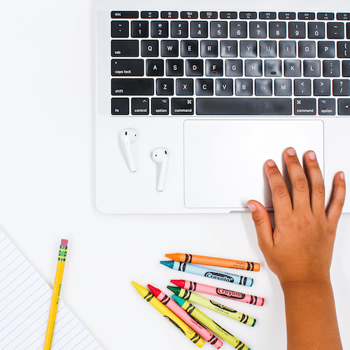 Flat lay image of desktop with crayons, AirPods and a child's hand on macbook laptop